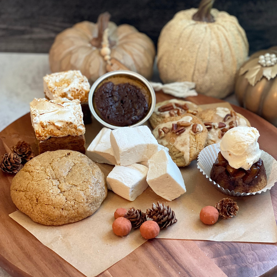 Desserts (including cookies, marshmallows, blondies, and other sweets) displayed on a wooden stand with decorative pumpkins in the backround.